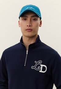 Jovem a usar um boné azul e um suéter azul-marinho com fecho até ao peito e logótipo "LD" no peito, voltado para a frente contra um fundo simples.