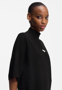 Zwarte gebreide top met een hoge kraag, wijde mouwen en een klein wit logo op de borst. Gladde textuur en modern ontwerp.