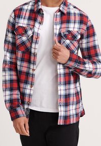 Rød, hvid og blå ternet flannelskjorte med to brystlommer, knapper ned foran og en lige kant. Båret over en hvid T-shirt.