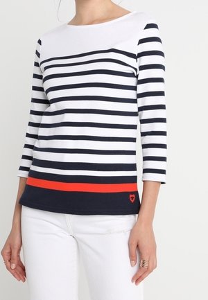 Kvinde iført en hvid langærmet topp med sorte horisontale striber og en rød stribe nær kanten, kombineret med hvide bukser.