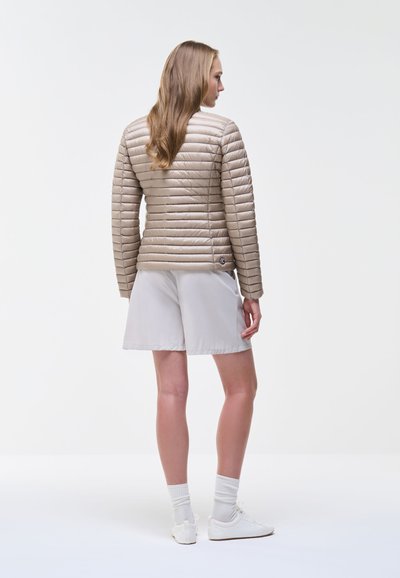 Femme aux longs cheveux, vêtue d'une veste matelassée beige, d'un short clair, de chaussettes blanches et de baskets blanches, debout de dos sur un fond uni.