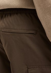 Κοντινή λήψη σκούρων καφέ παντελονιών με ελαστική μέση και λεπτομέρεια με φερμουάρ σε τσέπη, σε συνδυασμό με μπεζ πλεκτό πουλόβερ.