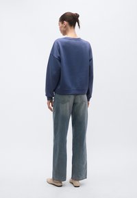 Μωβ φαρδιά φούτερ με φαρδιά εφαρμογή και χαλαρή γραμμή ώμων· συνδυασμένο με ανοιχτό γκρι τζιν παντελόνι φαρδιάς γραμμής και μπεζ παπούτσια τύπου slip-on.