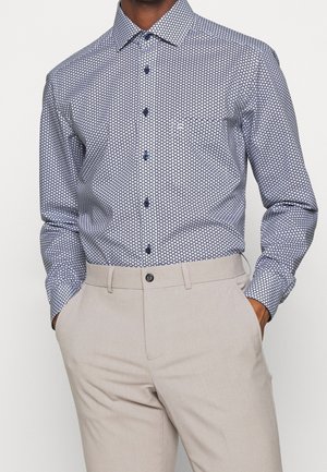 Homme portant une chemise bleue à motifs à manches longues boutonnée, rentrée dans un pantalon beige clair, les mains dans les poches.