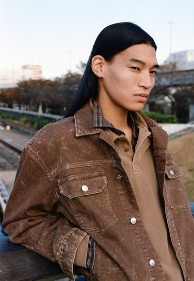 Young person with long black hair wearing a brown jacket and beige shirt stands outdoors leaning on a railing by a park pathway.