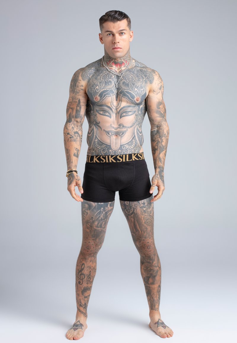 Homme musclé portant un short noir, recouvert de tatouages détaillés, dont un grand motif de visage sur la poitrine et divers tatouages aux manches et aux jambes.