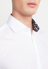 Camicia bianca con collare a motivo floreale nero. Tessuto morbido, design aderente e forma del colletto classica.