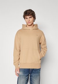 Sudadera con capucha beige de tejido suave, con un bolsillo frontal y puños acanalados. Presenta un logotipo sutil en el pecho. Se combina con jeans azules.