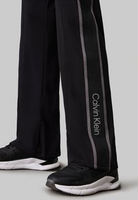 Pantaloni sportivi neri con strisce grigie, con logo Calvin Klein. Realizzati in materiale liscio e elastico. Abbinati a scarpe da ginnastica nere.