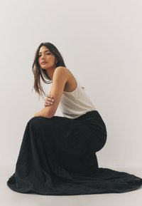 Czarna długa spódnica maxi o fakturowanym, zwiewnym kroju w połączeniu z lekkim, prążkowanym, bezrękawnym topem. Modelka siedzi na tle gładkiego tła.