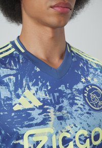 Maglia da calcio blu con accenti gialli, caratterizzata da un motivo astratto testurizzato, colletto a V e loghi Adidas e Ajax sul petto.