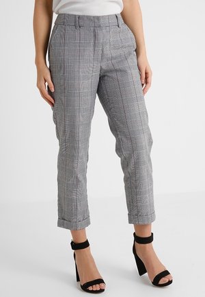 Vrouw draagt grijze geruite cropped broek met omslagen en zwarte enkellaars sandalen met hoge hak en enkelbandje, staand tegen een effen achtergrond.