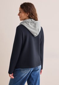 Merivartijan collegepaita harmaalla hupulla, jossa on sileä pinta ja pitkät hihat. Takana on rento istuvuus ja epämuodollinen muotoilu.