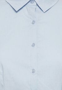 Svijetloplena košulja kratkih rukava s ovratnikom, glatke teksture i gumbima sprijeda koji su od prozirne plastike, s ukrasnim šavovima.