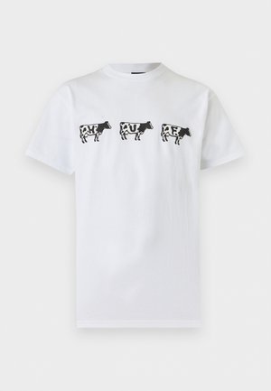 Jeune homme aux cheveux tressés porte un t-shirt blanc avec trois silhouettes noires de vaches avec des lettres à l'intérieur, associé à un pantalon gris clair.