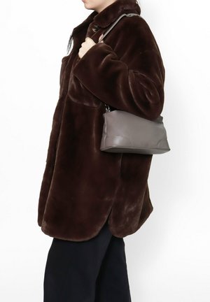 Persoon draagt een donkerbruine imitatiebonten jas en houdt een kleine grijze leren schoudertas vast tegen een effen witte achtergrond.