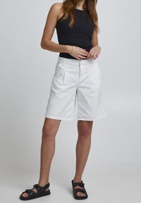 Hvide shorts med frontfolder, lynlås og sidelommer, parret med en sort ærmeløs top og sorte sandaler. Glat stoftekstur.