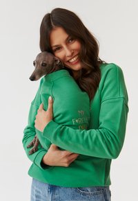 Groene sweatshirt met geribbelde boorden, met de speelse tekst "MIJN VOOR ALTIJD DATUM." Een klein hondje wordt nauwkeurig vastgehouden, wat een zachte, gezellige textuur toont.