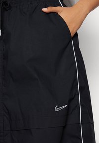 Svart vindjacka med dragsko i midjan, sidoficka, vit kantning och en liten broderad Nike-logga på nedre högra framsidan.