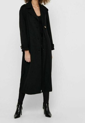 Frau, die einen langen schwarzen Trenchcoat, schwarze Hosen und schwarze hochhackige Stiefeletten trägt, steht vor einem schlichten Hintergrund.