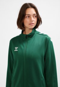 Grön sportjacka med dragkedja och hög krage, tillverkad av ett slätt tyg. Har vit logotyp och randiga detaljer på axlarna.