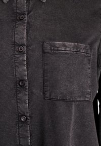 Schwarzes Knopfleib-Hemd mit glatter Textur, ausgestattet mit einer aufgesetzten Tasche auf der linken Seite und schwarzen Knöpfen entlang der vorderen Knopfleiste.