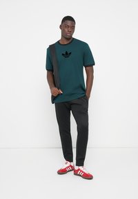 Grøn bomulds T-shirt med sort logo, sort kant, sorte bukser og rød-hvid-stribede træningssko. Modellen holder en sort skuldertaske.