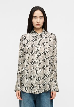 Femme aux longs cheveux foncés portant un chemisier à manches longues imprimé serpent noir et blanc et un jean bleu, debout devant un fond uni.