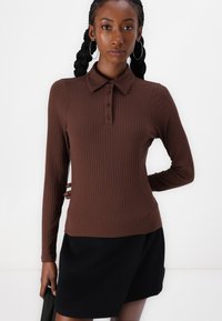 Brown ribbed long-sleeve polo top with a collared neckline and three-button placket, paired with a black skirt. Smooth texture and fitted design.