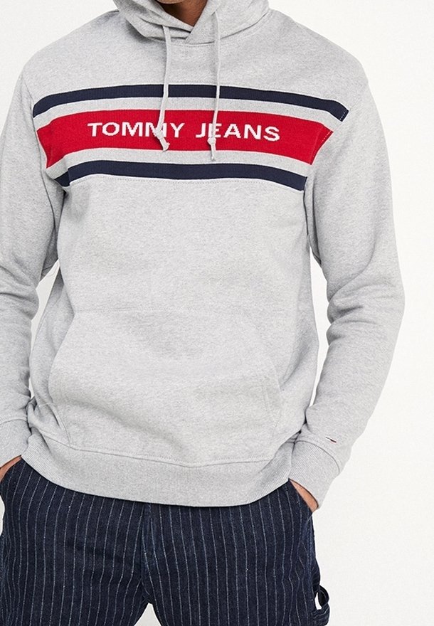 Sweat à capuche gris avec des bandes rouges, blanches et bleu marine sur la poitrine, portant le texte "TOMMY JEANS", associé à un pantalon bleu marine à fines rayures.