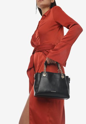 Borsa in pelle nera con accenti in catena argentata, forma arrotondata e base piatta. Il modello indossa un vestito rosso con maniche lunghe e una cintura in vita.