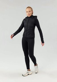 Svart hettejakke med glidelås, quiltede paneler, lange ermer, sammen med svarte leggings og hvite treningssko. Glatt stofftekstur.