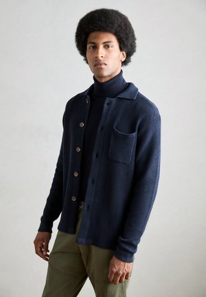 Marineblå cardigan med struktureret strik, knaplukning foran og en brystlomme. Bruges over en matchende turtle neck og parrer med khakibukser.