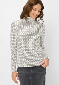 Langærmet turtleneck-top i creme med sort geometrisk mønster, med en tætsiddende silhuet og blødt, strækbart stof. Glat tekstur.