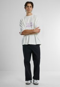 Hvit oversized t-skjorte med "CIAO AMORE" i lilla tekst, paret med mørke bukser og grå-hvite sneakers. Glatt bomullsmateriale.