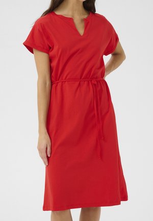 Kobieta w jaskrawoczerwonej sukience za kolano z krótkimi rękawami, dekoltem w kształcie litery V i cienkim paskiem w talii, stojąca na jednolitym tle.