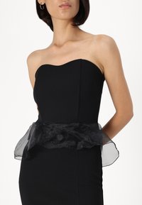 Vestido preto sem alças, com um corpete justo e sobreposição preta transparente e com babados na cintura. Tecido liso; linhas limpas; silhueta elegante.