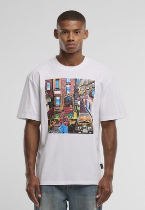 Tânăr purtând un tricou alb cu imprimeu colorat de artă urbană stradală, ce prezintă clădiri, oameni și mașini parcate.