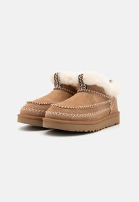 Zapatillas de ante marrón con forro de piel de oveja blanca, costuras decorativas y suelas de goma. Punta redondeada y diseño bajo para un uso fácil.