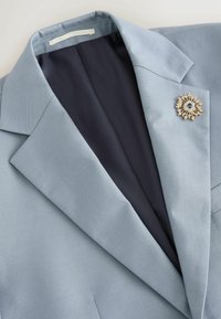 Blazer bleu clair avec une doublure intérieure foncée et une broche décorative dorée et bleue épinglée sur le revers gauche.