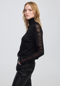 Vrouw in een zwarte coltrui met doorzichtige, gerimpelde mouwen en zwarte leren broek, staand in profiel tegen een effen lichtgrijze achtergrond.