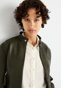 Chaqueta bomber de cuero verde oscuro con cierre de cremallera en la parte frontal, cierre a presión y bolsillos laterales, llevada sobre una blusa blanca con detalles de encaje.
