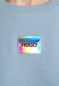 Maglietta blu in maglia con una toppa quadrata olografica che riporta il testo "HUGO" in nero, centrato sul petto.
