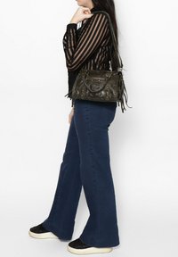 Zwarte leren handtas met zilveren studs, ritsvak en franjes. Model draagt een doorzichtige zwart gestreepte top en blauwe flared jeans, met zwarte sneakers.