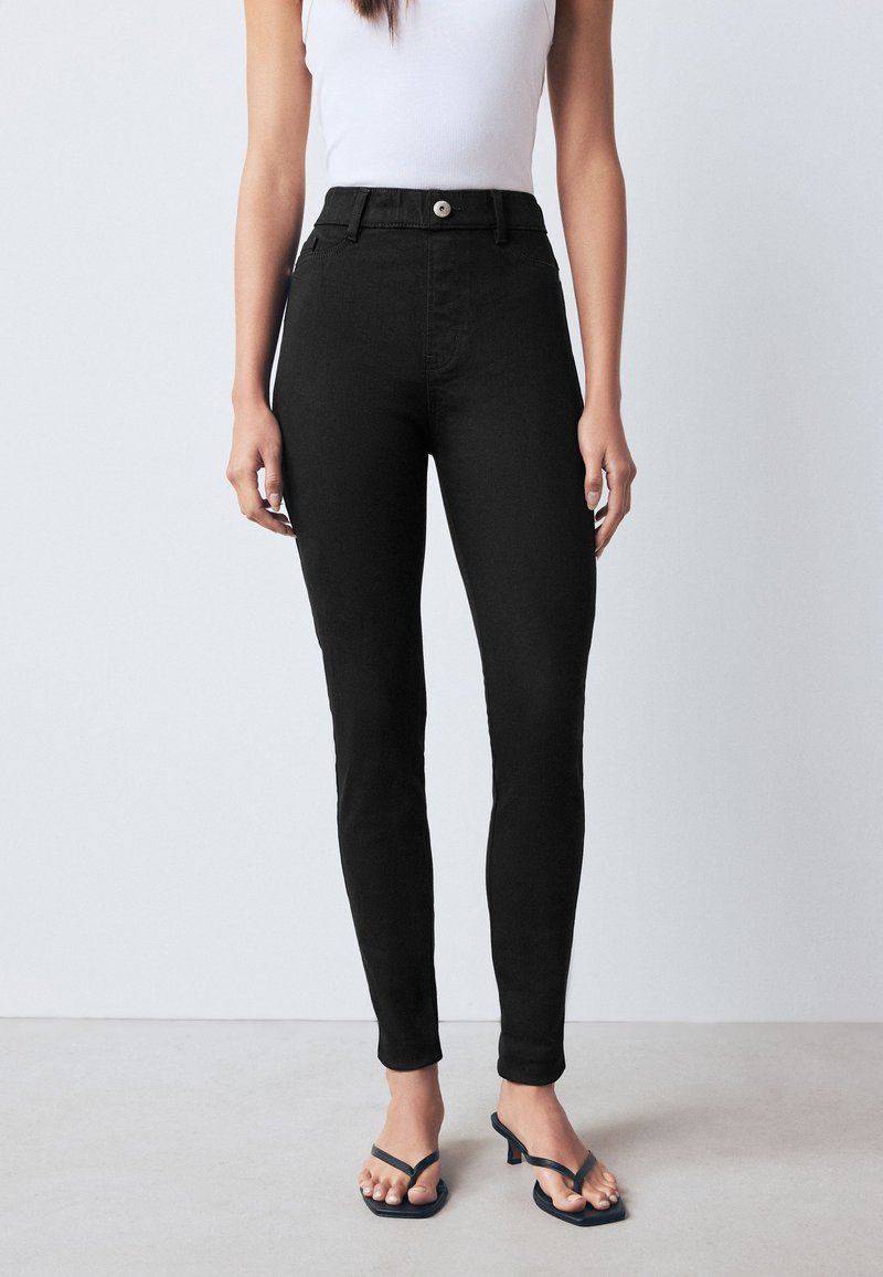 Vrouw die zwarte skinny jeans met hoge taille, knoop en rits draagt, gecombineerd met zwarte sandalen met hak en een wit mouwloos top.