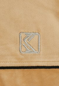 Bež antilop tkanina z mehko teksturo, ki vsebuje srebrni vezen logotip v pravokotnikasti obliki z nagibno črto pod njim.