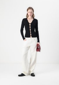 Maglietta nera a maniche lunghe con scollatura a V e bottoni decorativi, abbinata a pantaloni bianchi larghi e una piccola borsa rossa. Le scarpe nere completano il look.
