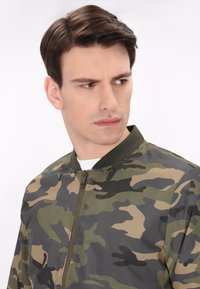 Camouflagejack in groen, bruin en zwart. Beschikt over een ritssluiting aan de voorkant, een geribde kraag en een lichtgewicht stof met een gladde textuur.
