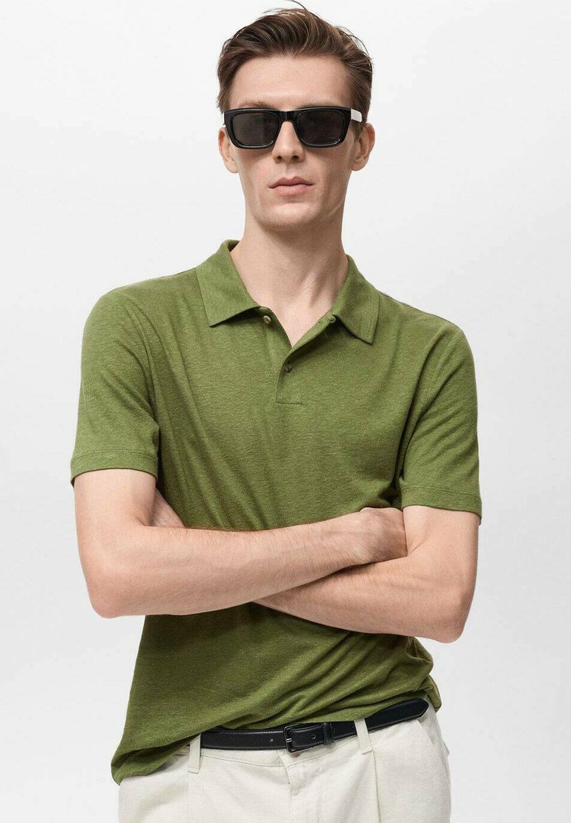 Zelená polo tričko na krátky rukáv s golierom a dvoma gombíkmi. Model má na sebe čierne slnečné okuliare, krémové nohavice a čierny opasok.