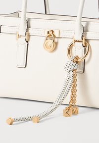 Witte leren handtas met gouden hardware, voorzien van een rond logo, een decoratieve ketting en een polkadot franjes-detail.
